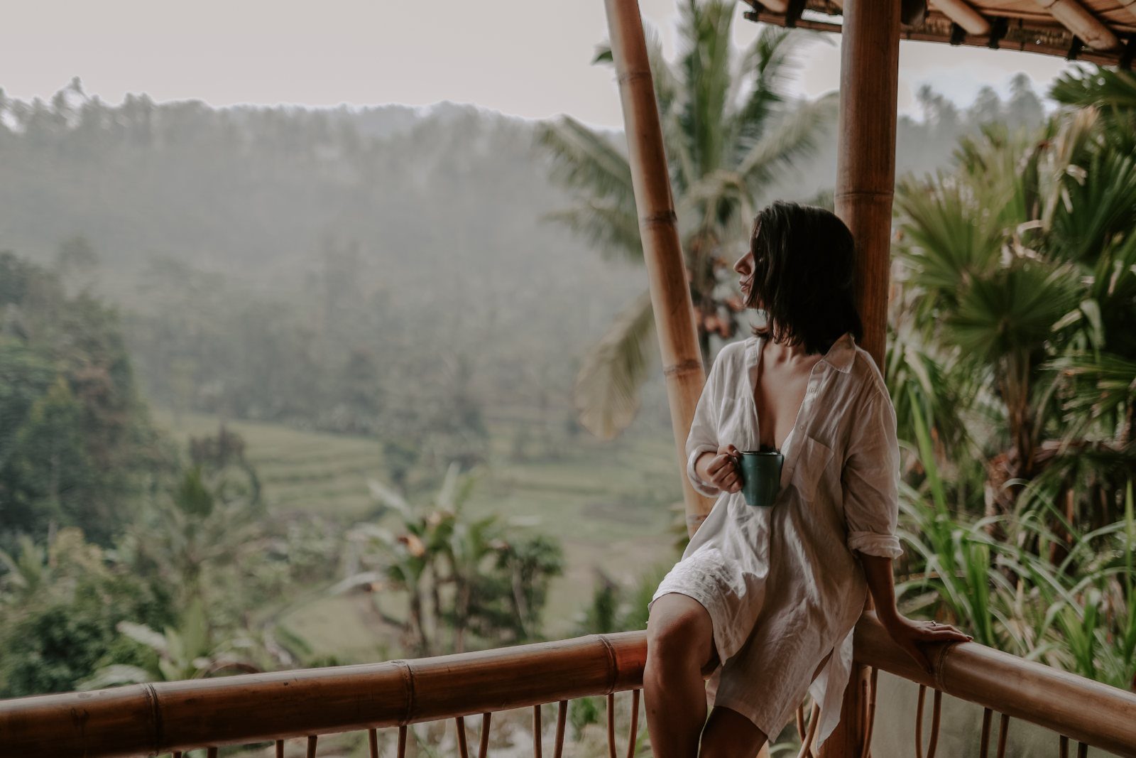 A woman with a mug on a bamboo balcony gazes over misty jungle, a calm pause to ask, is psychedelic therapy right for me.
