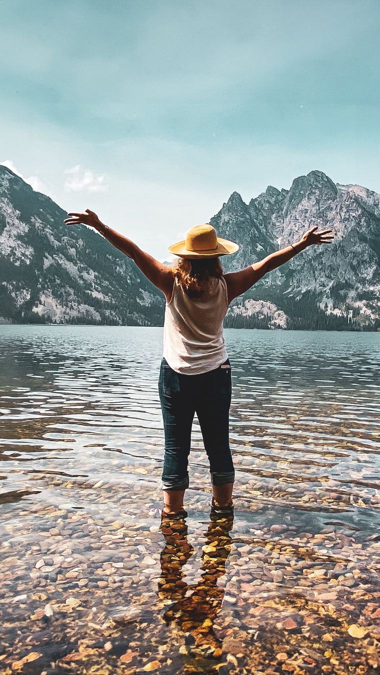 A woman stands in a clear alpine lake, arms open toward the mountains, seen from behind.