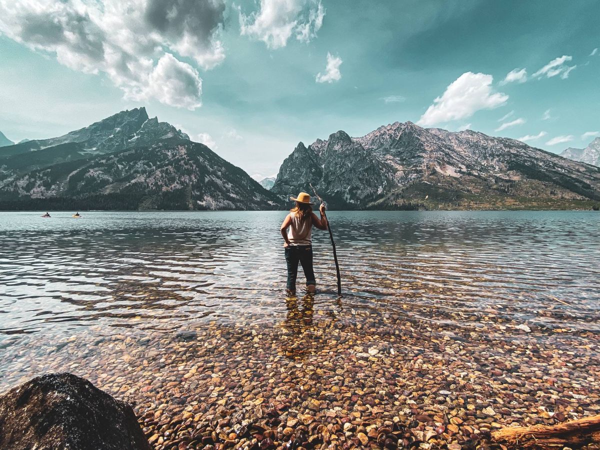 A person in a sun hat stands in a clear mountain lake with a walking stick, facing rugged peaks